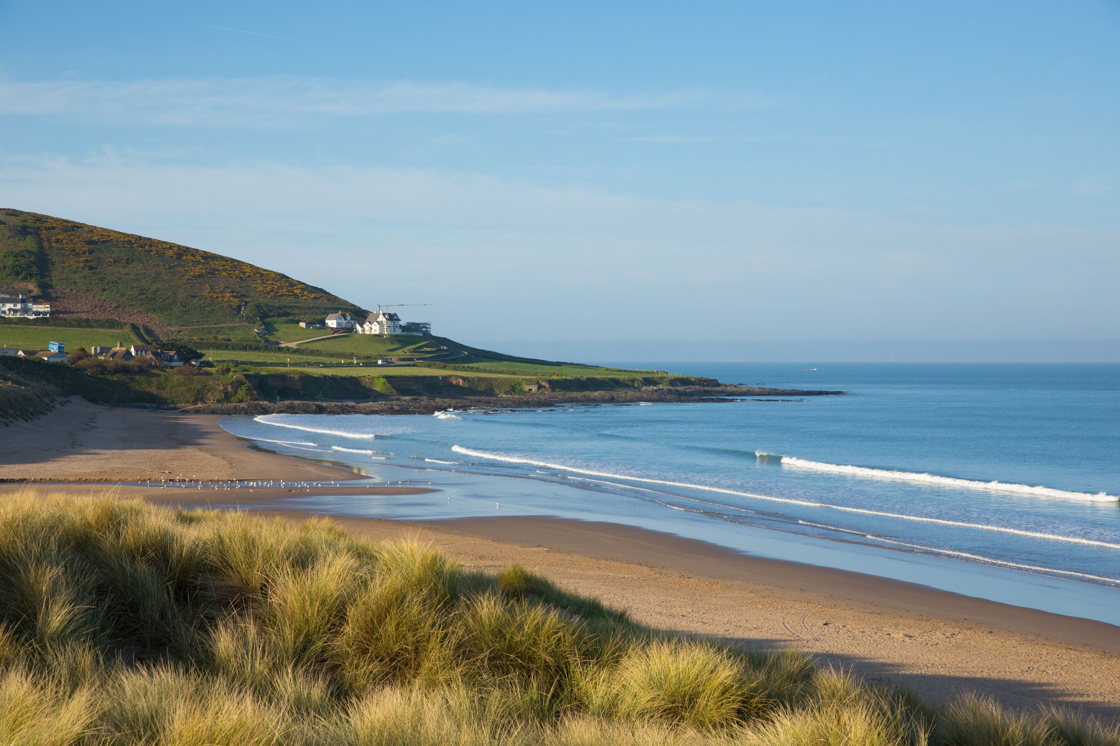 A coastal landscape with a sandy beach, grassy dunes, and a hill with houses.