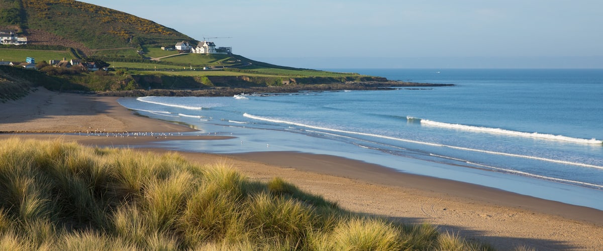A coastal landscape with a sandy beach, grassy dunes, and a hill with houses.