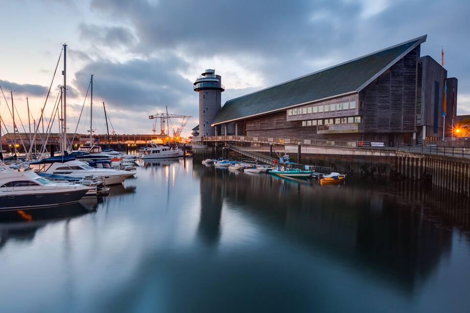 Un port esportiu amb vaixells amarrats, un far i un edifici modern amb teulada inclinada.