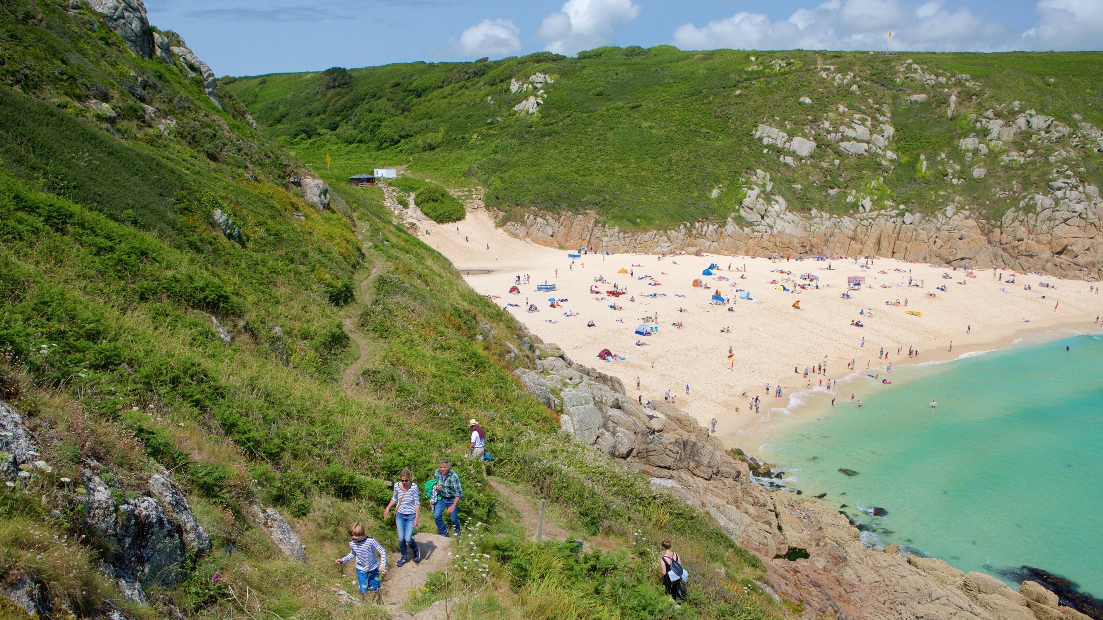 A coastal beach with people sunbathing and swimming, surrounded by green hills and rocky cliffs.