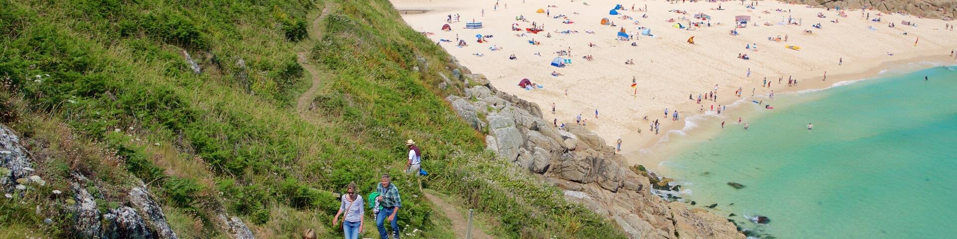 A coastal beach with people sunbathing and swimming, surrounded by green hills and rocky cliffs.