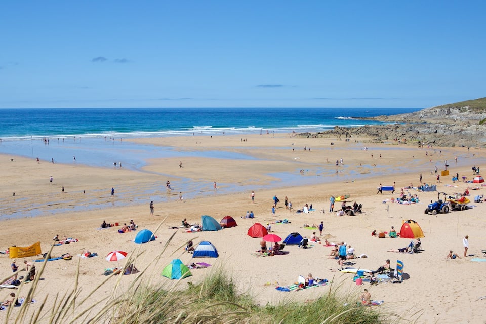 Una playa concurrida donde gente se toma el sol, hay tiendas de campaña y una colina rocosa al fondo.