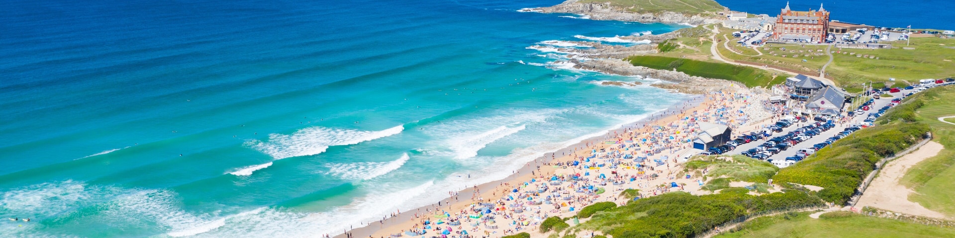 Uma praia costeira com uma grande multidão, um edifício histórico e um céu azul claro.