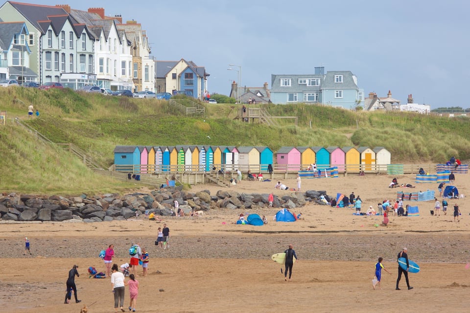 A beach with colorful beach huts, people walking, and houses in the background.