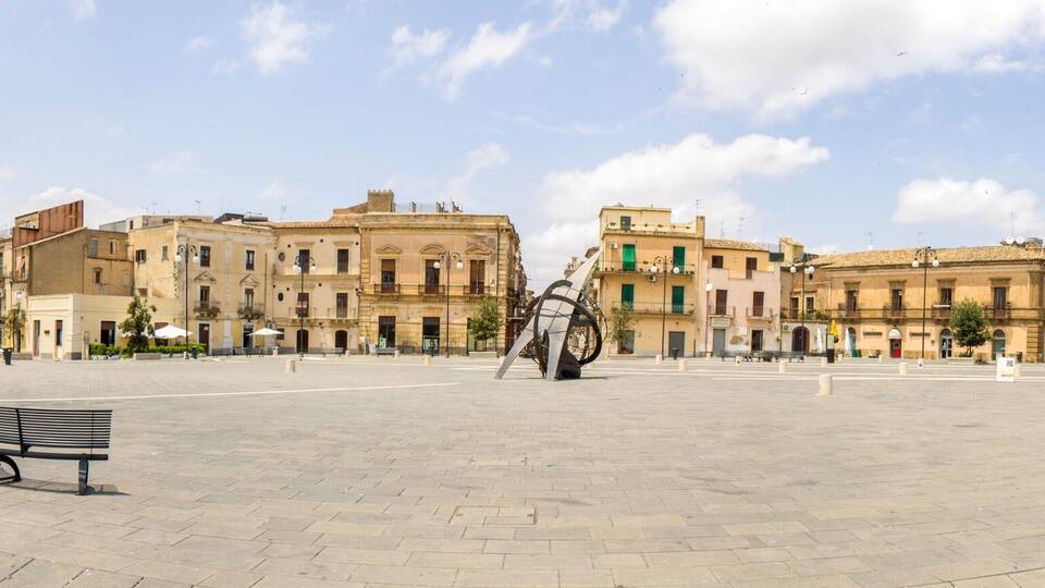 A spacious square with historic buildings, a prominent clock tower, and a statue in the center.