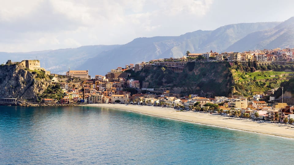 A coastal town with a prominent castle on a cliff, a beach, and mountains in the background.