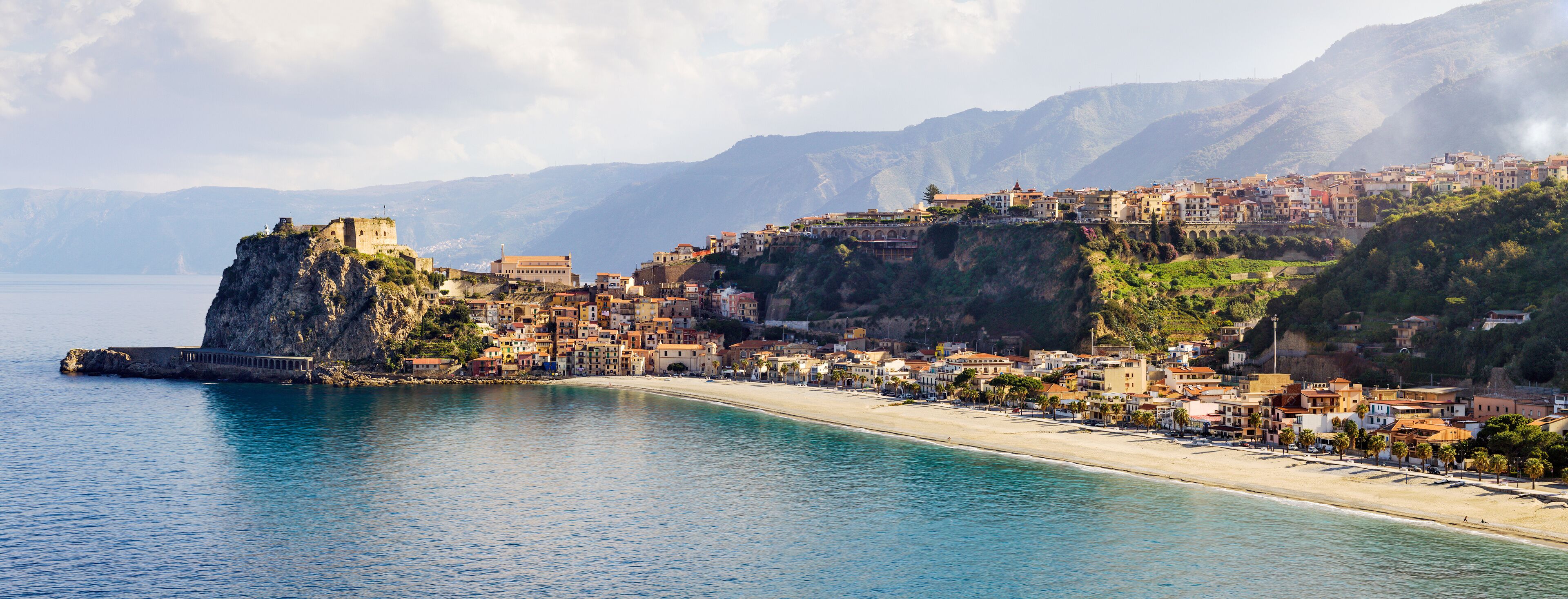 A coastal town with a prominent castle on a cliff, a beach, and mountains in the background.