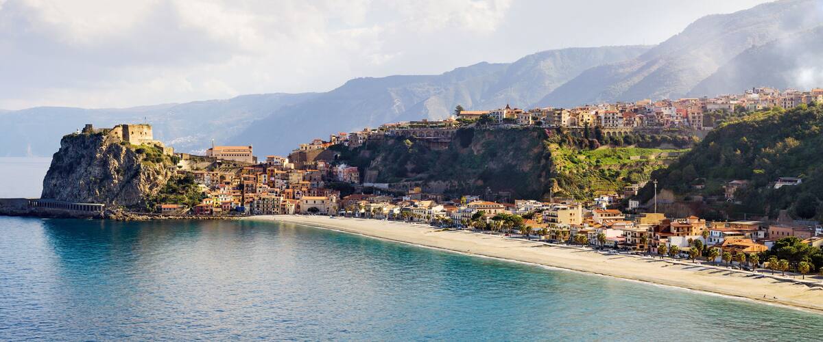 A coastal town with a prominent castle on a cliff, a beach, and mountains in the background.