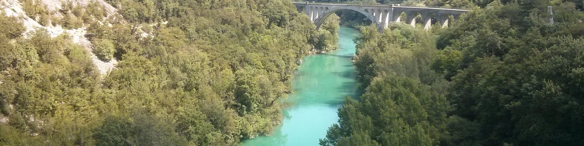 Un río turquesa serpentea por un valle boscoso, con un puente de piedra a lo lejos.