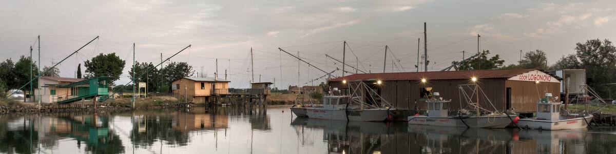 Ein ruhiger Hafen mit mehreren vertäuten Fischerbooten, einem kleinen Holzhaus und einem bewölkten Himmel.