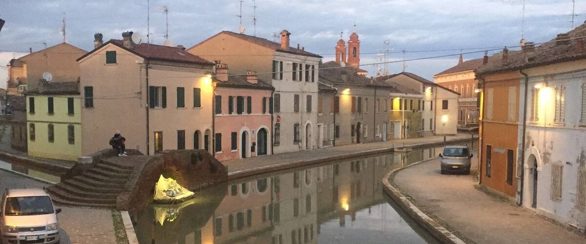 A canal with buildings reflecting on the water, a bridge, and a tower in the background.