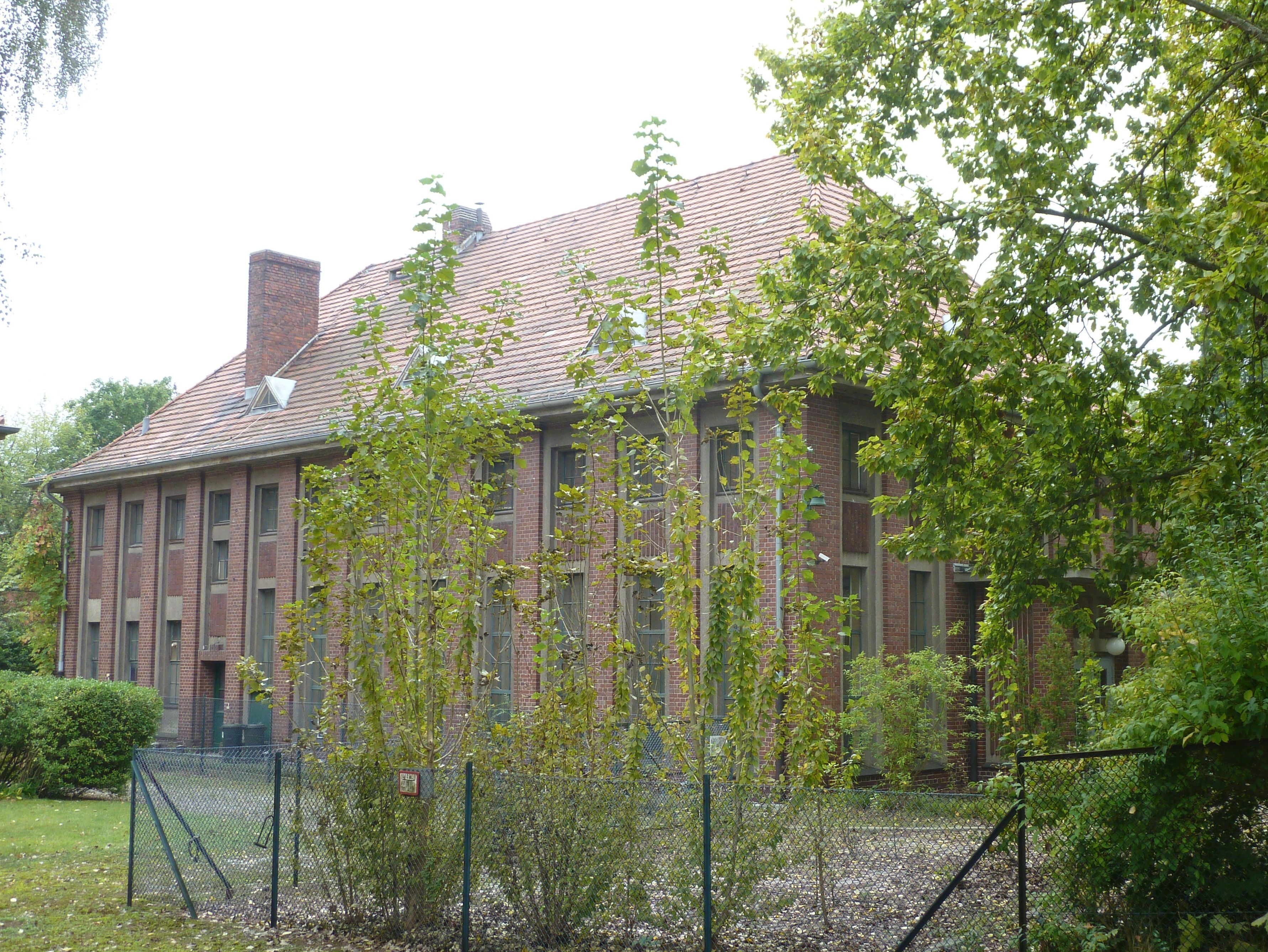 A brick building with a red-tiled roof, surrounded by trees and a chain-link fence.