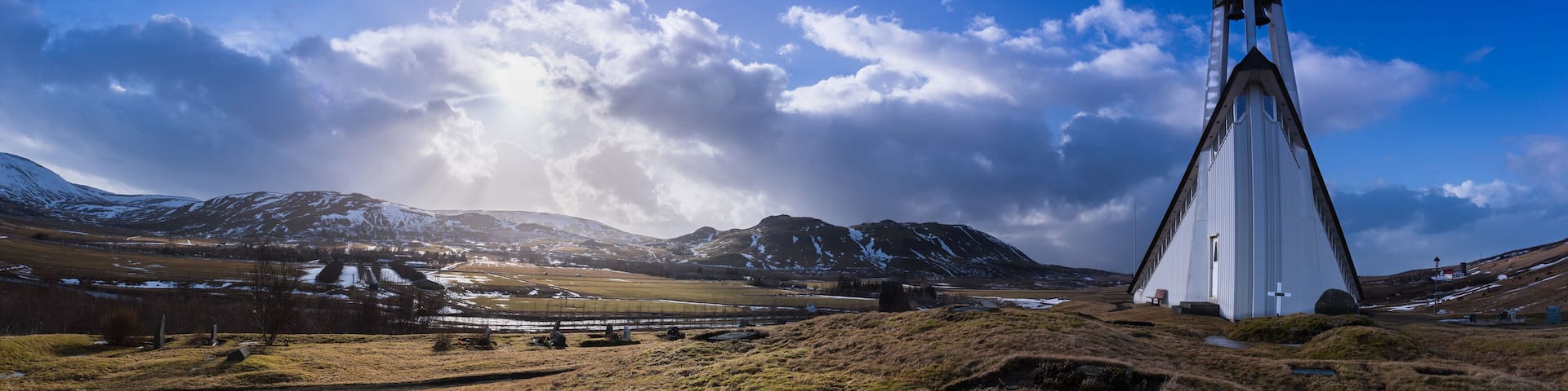 Panorama of the Mosfellsbaer church in Iceland at sunset with a cross, graves, green grass, mountains, clouds, and blue sky