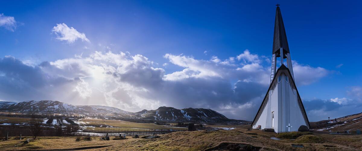 A church with a tall steeple in a rural landscape with mountains and snow.
