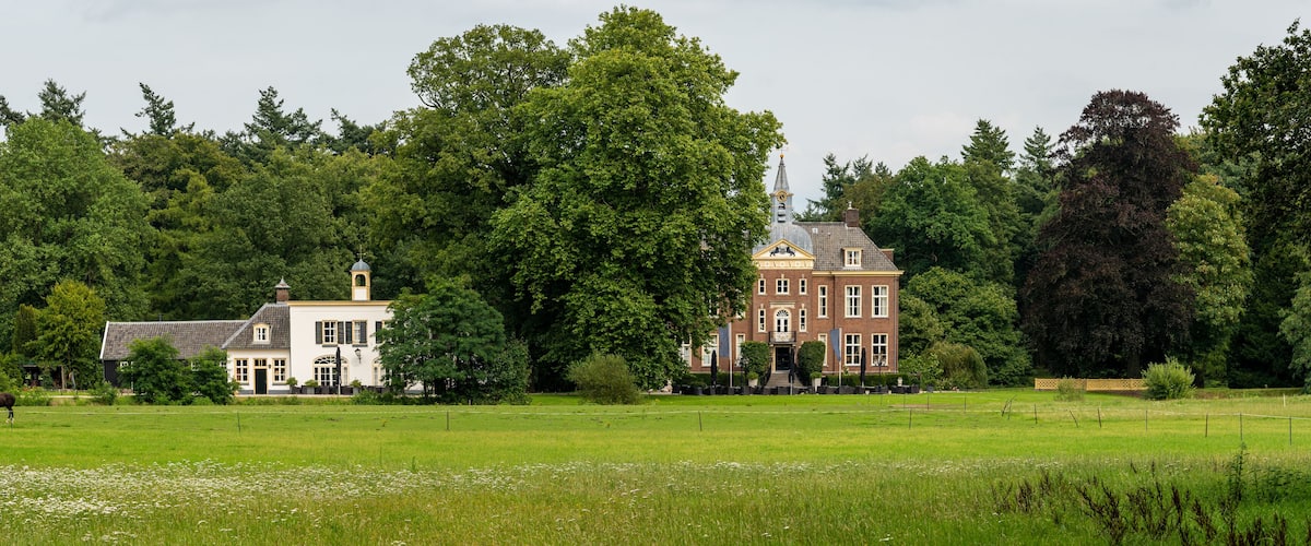 A large brick building with a tower, surrounded by lush green trees and a field with a horse grazing.