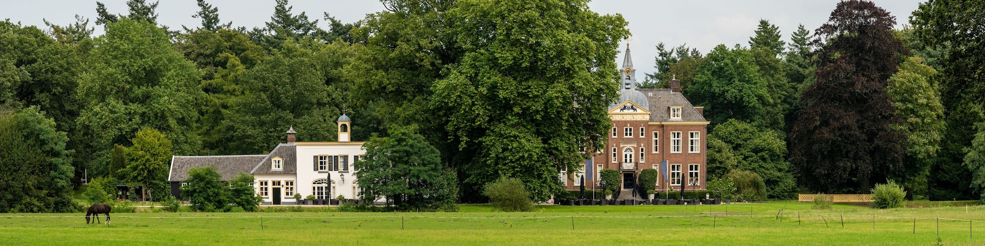 A large brick building with a tower, surrounded by lush green trees and a field with a horse grazing.