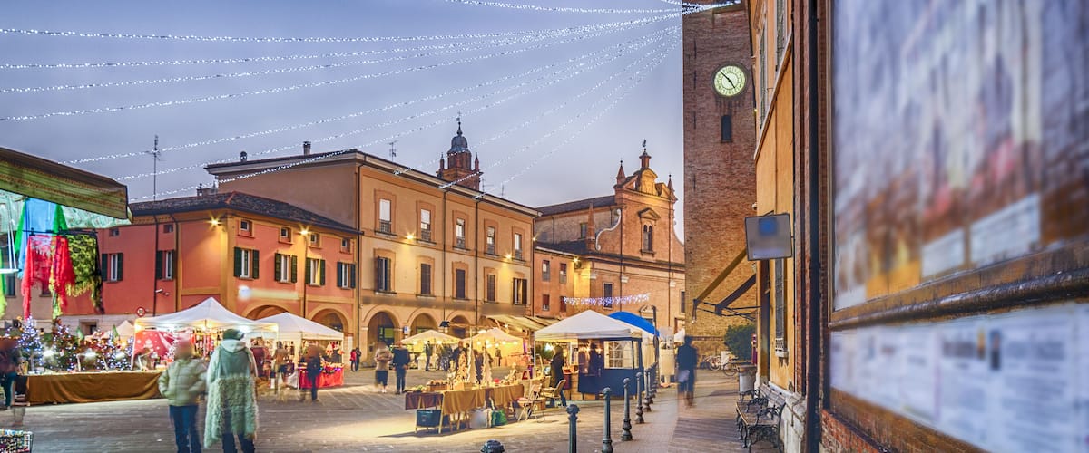 A historic square with market stalls, a clock tower, and string lights.