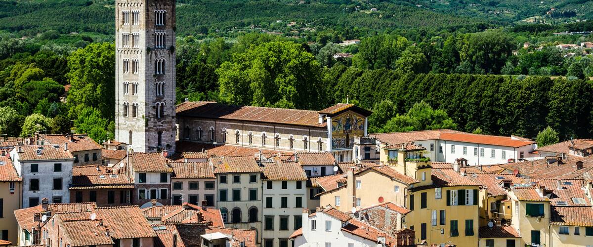 A town with a prominent tower, terracotta-roofed buildings, and lush green hills in the background.
