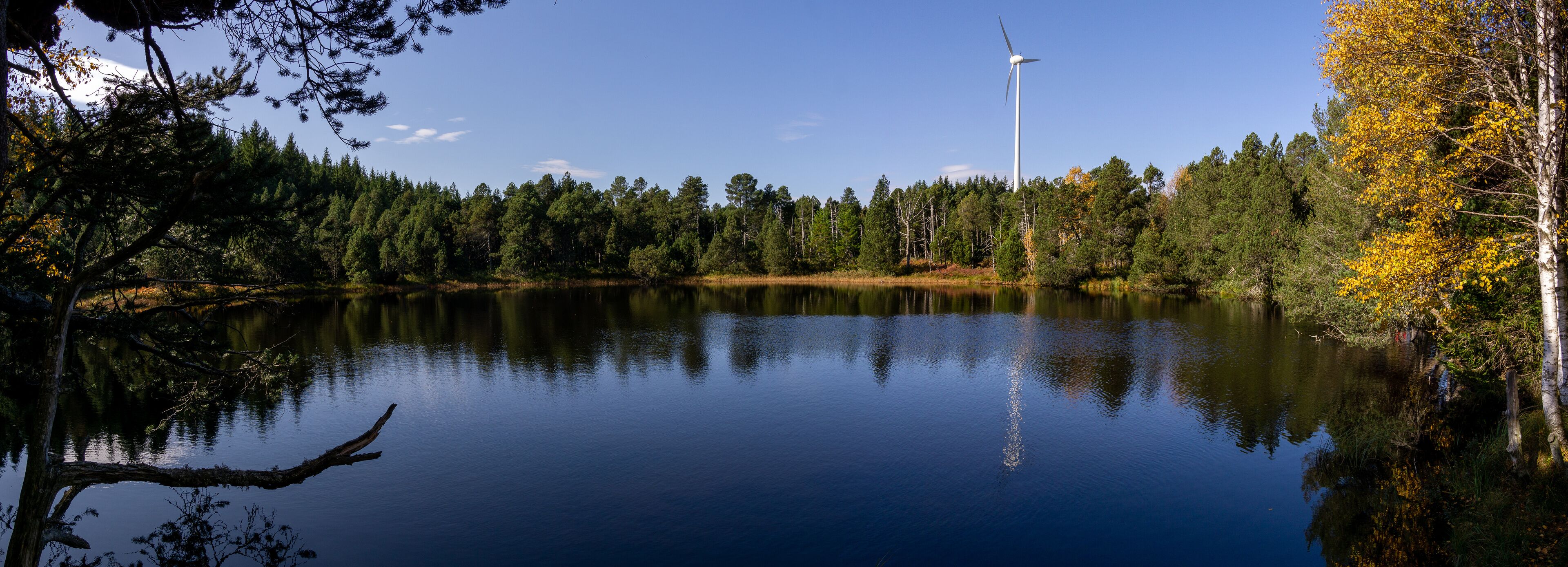 Un lago tranquilo con una turbina eólica a lo lejos y árboles de colores otoñales.