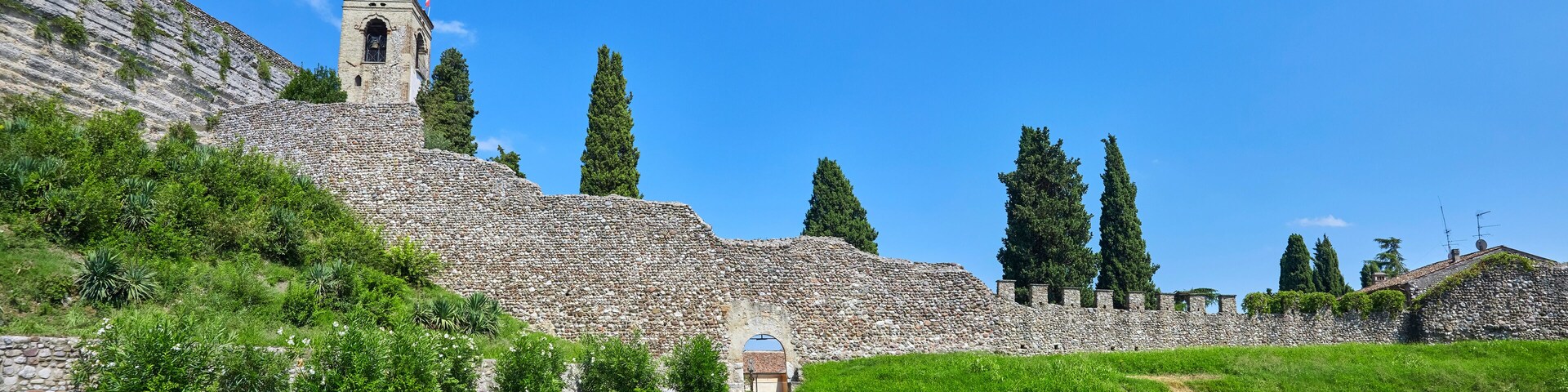 A historic stone wall with a tower and a gate, surrounded by greenery and tall trees.