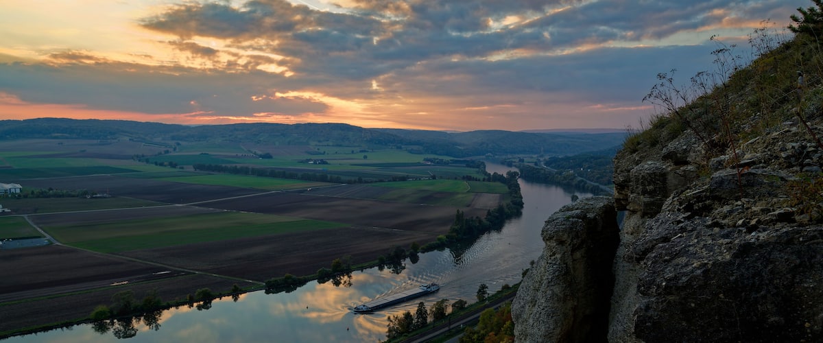 A river reflecting the sunset, surrounded by fields and a rocky cliff.