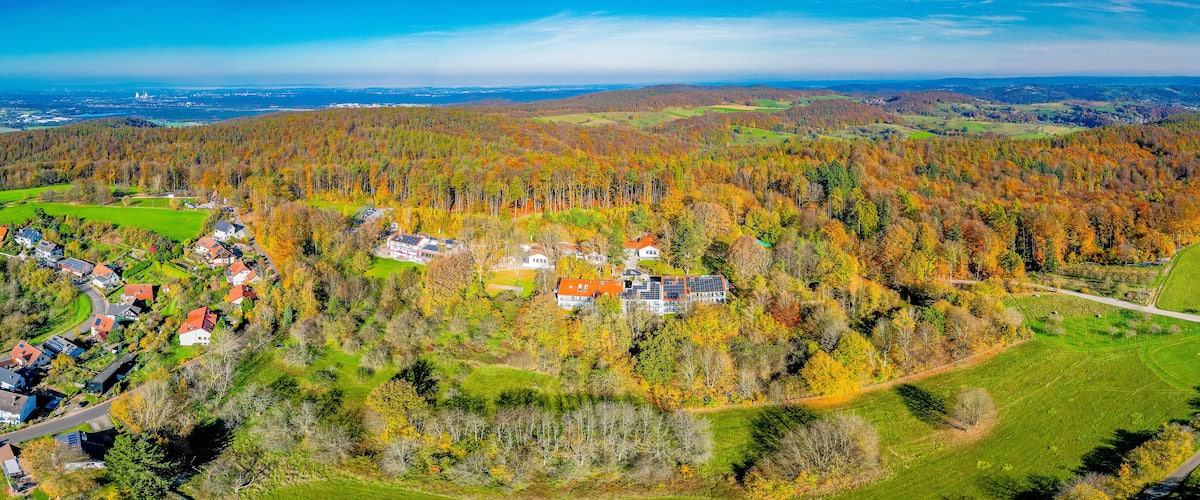 A village nestled in a forested valley with autumn foliage, surrounded by rolling hills and a distant view of a coastal city.