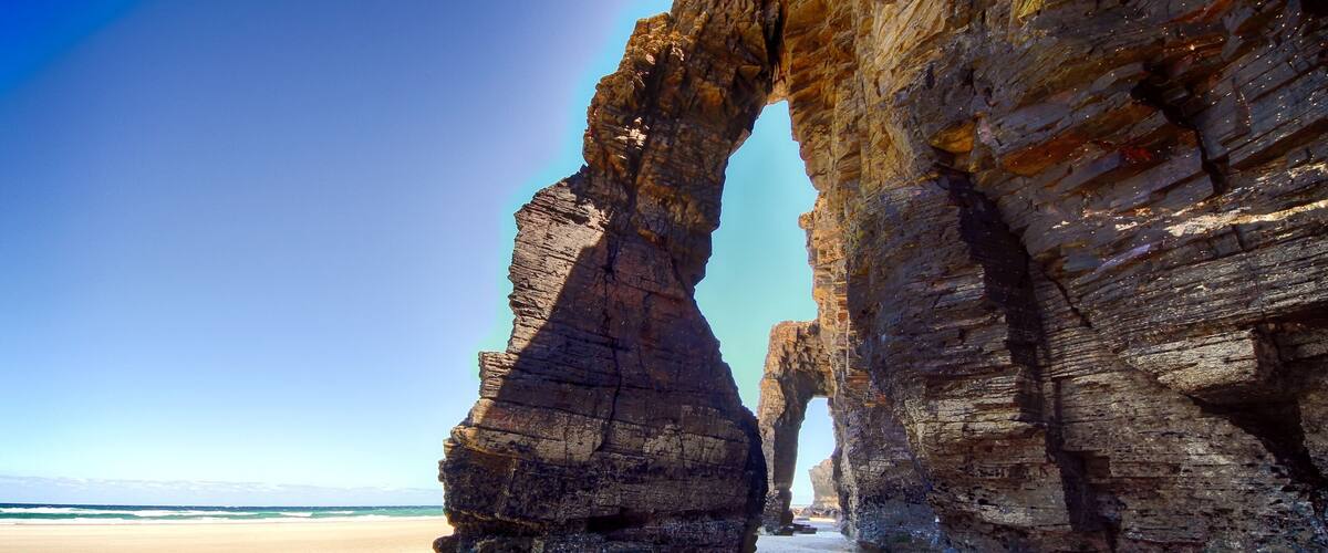 A natural rock arch formation on a beach with clear water and a sandy shore.