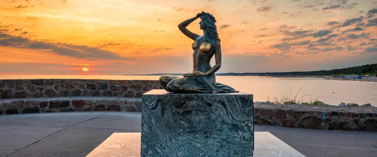 Una statua di una donna su un piedistallo, vicino alla riva al tramonto.