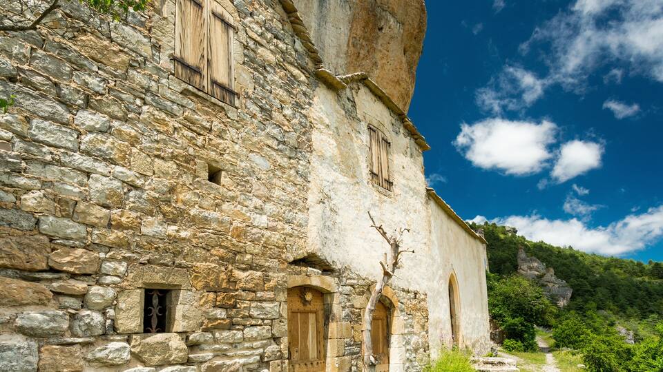 A stone building with wooden shutters and a path leading to a cliff.