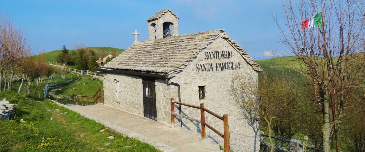 A small stone chapel named Santuario Santa Famiglia with a cross on top, surrounded by green hills and a clear sky.