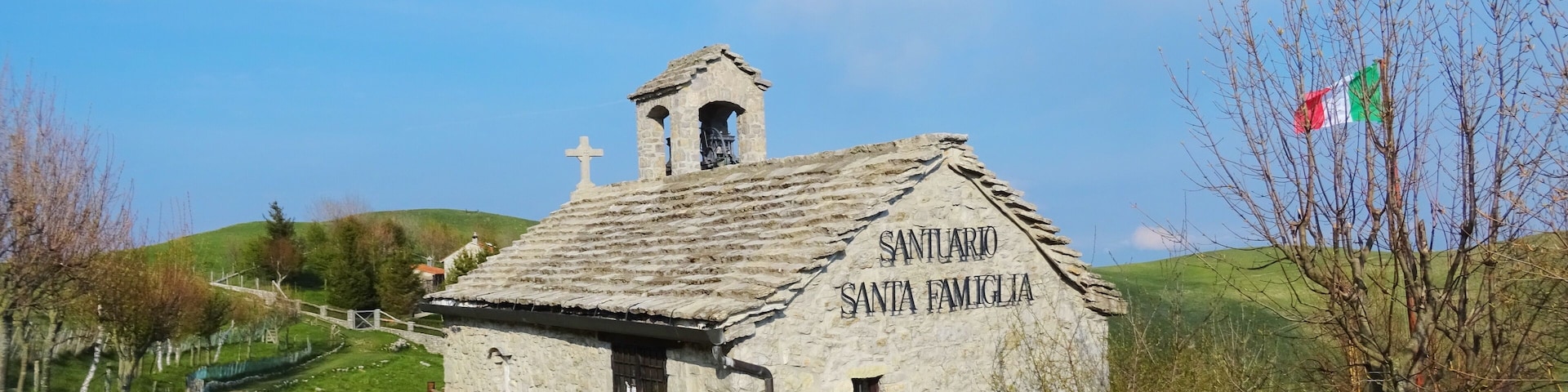 A small stone chapel named Santuario Santa Famiglia with a cross on top, surrounded by green hills and a clear sky.
