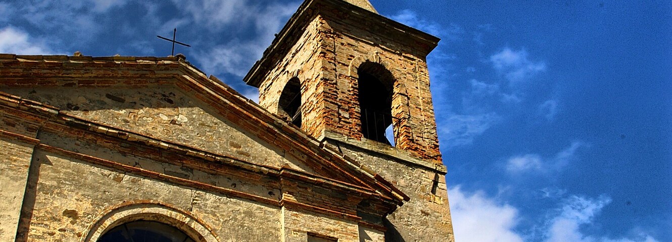 Una iglesia con un campanario y una cruz en la parte superior, rodeada de árboles y un cielo parcialmente nublado.