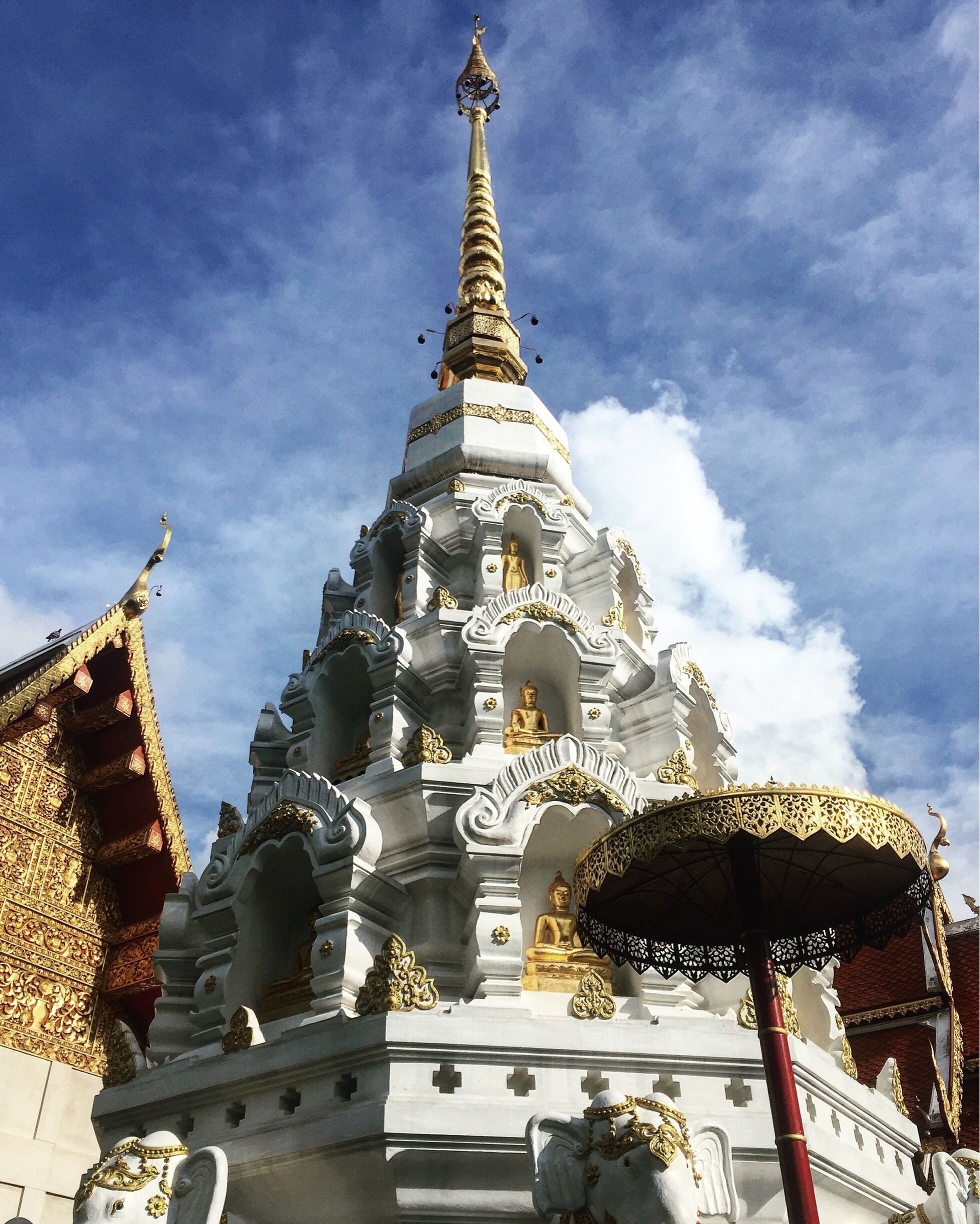 A white and gold stupa with intricate carvings and a golden spire, surrounded by smaller golden statues and a red pillar.