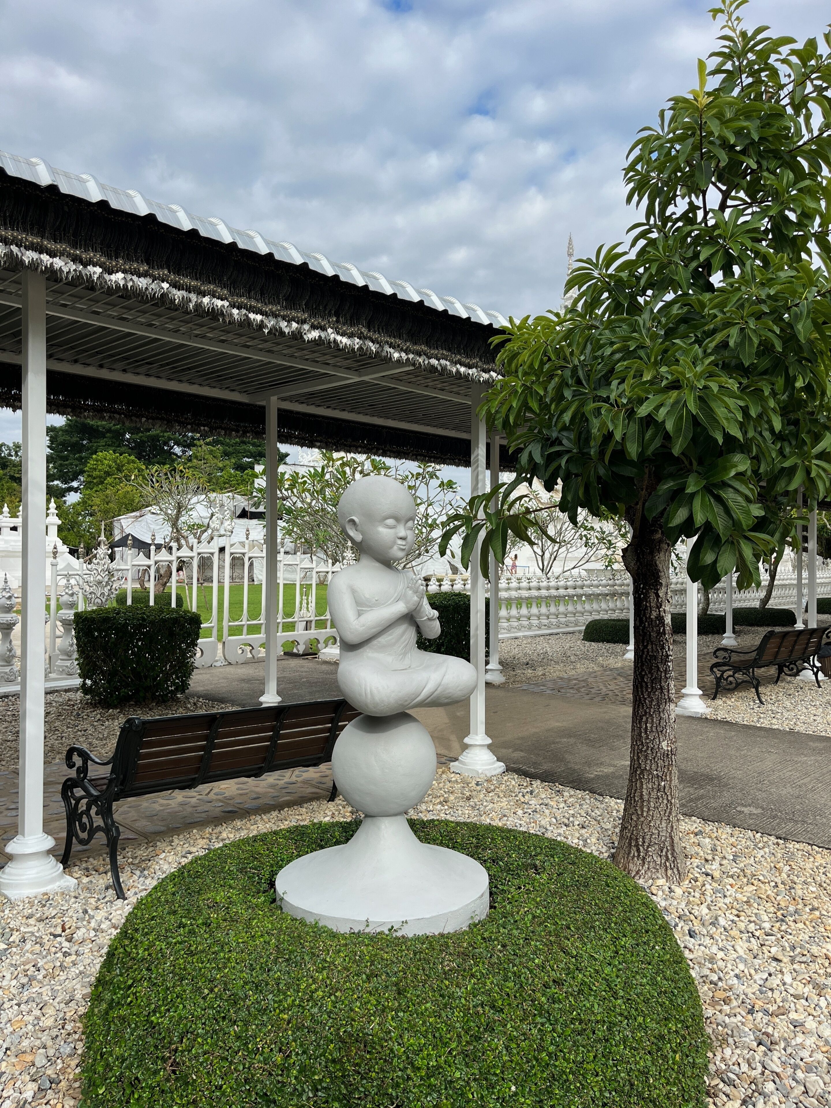 Escultura de uma criança em pedestal em parque, com árvore, banco e pavilhão.