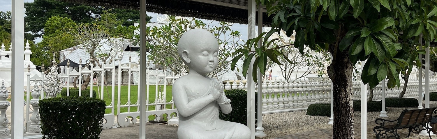 Escultura de uma criança em pedestal em parque, com árvore, banco e pavilhão.