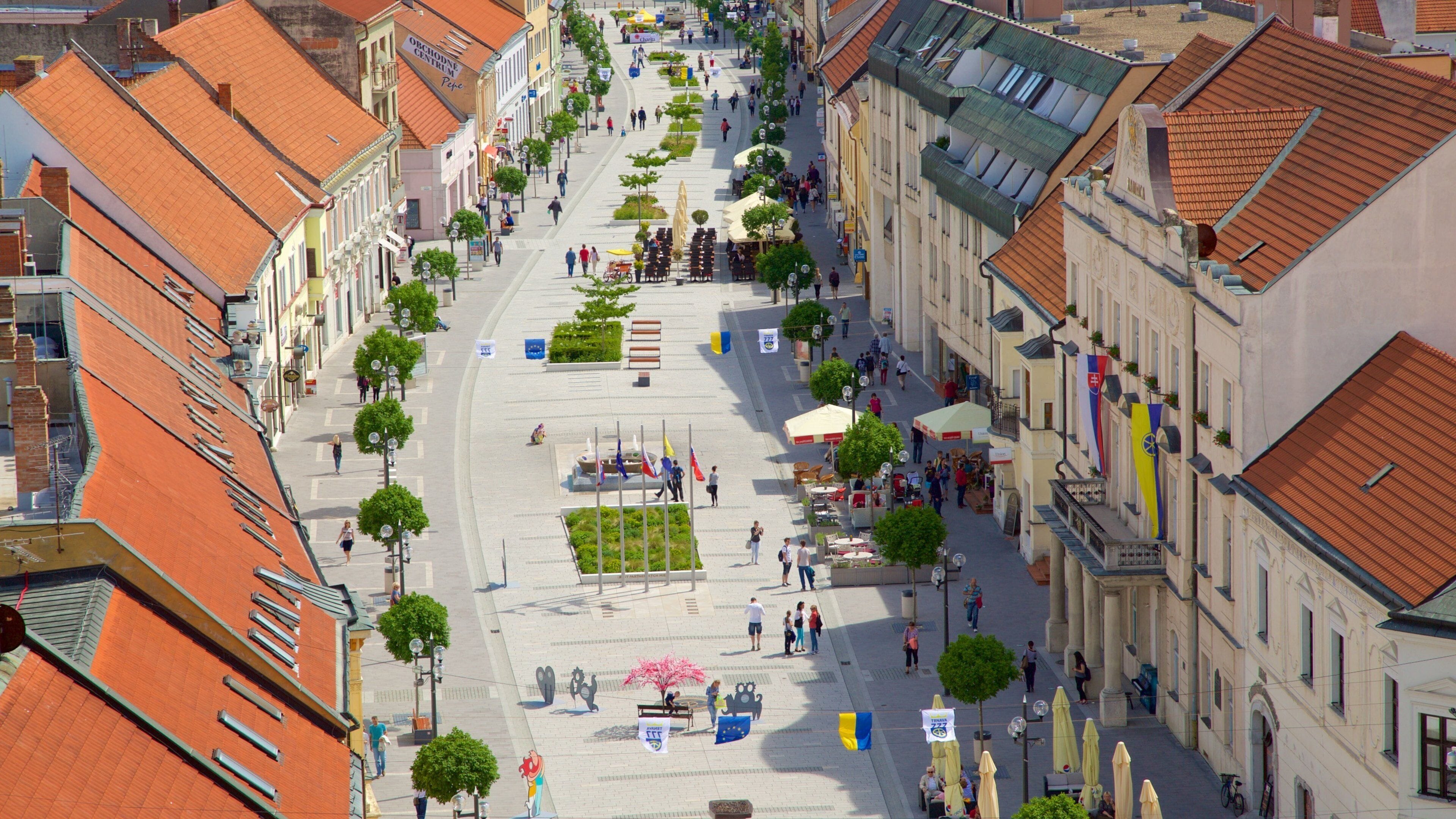 A wide street lined with buildings, trees, and outdoor seating areas.