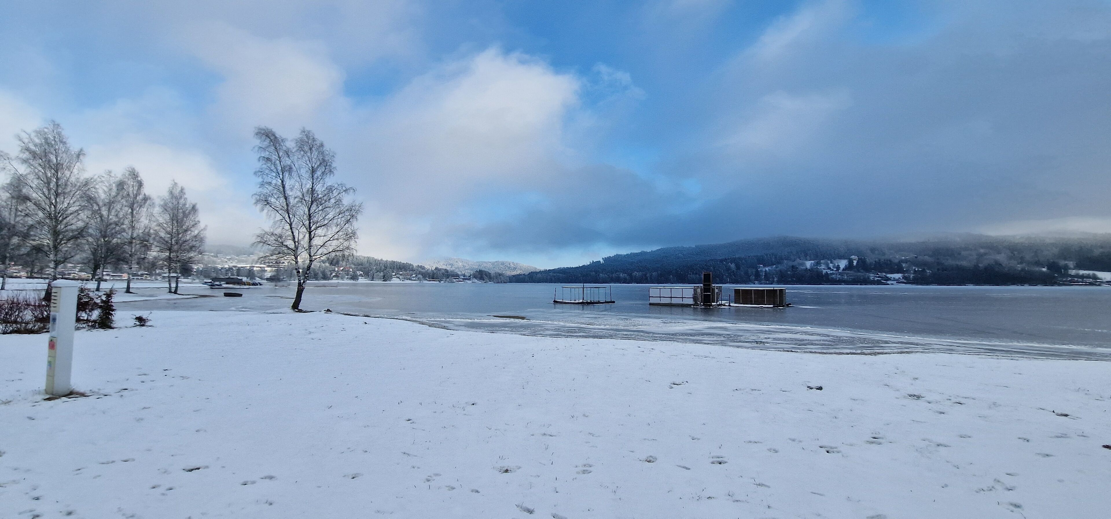 A snow-covered landscape with a frozen lake, leafless trees, and distant mountains under a cloudy sky.