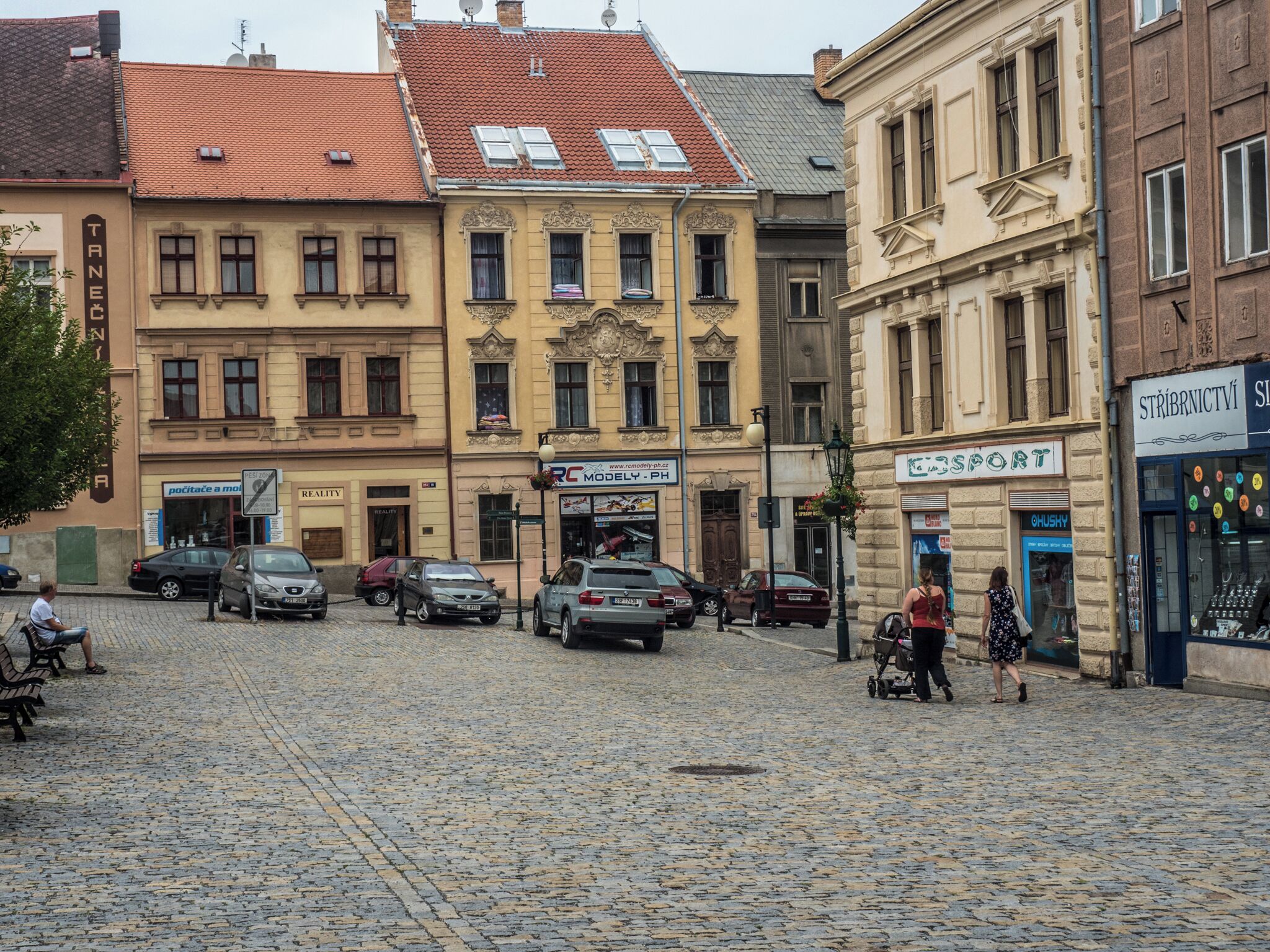 Una calle adoquinada con edificios históricos, coches estacionados y una mujer empujando un cochecito.