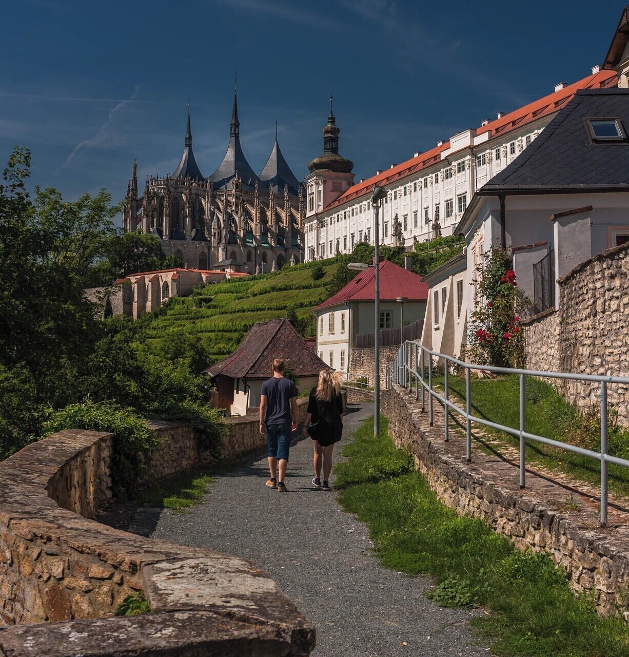 A pathway leading to a historic building with multiple spires, surrounded by lush greenery and stone walls.