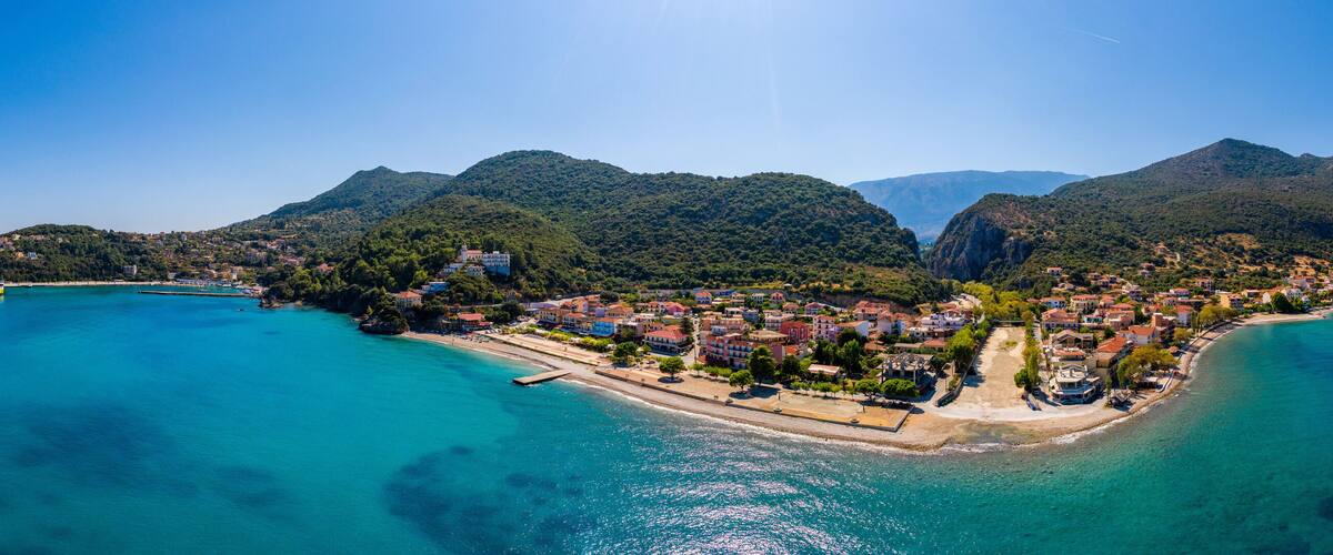 An aerial view of a coastal town with a clear blue sea, surrounded by lush green hills and mountains.