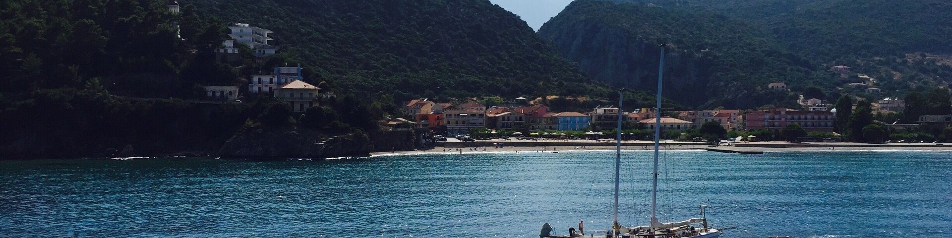 A sailboat on a calm sea with a coastal town and mountains in the background.