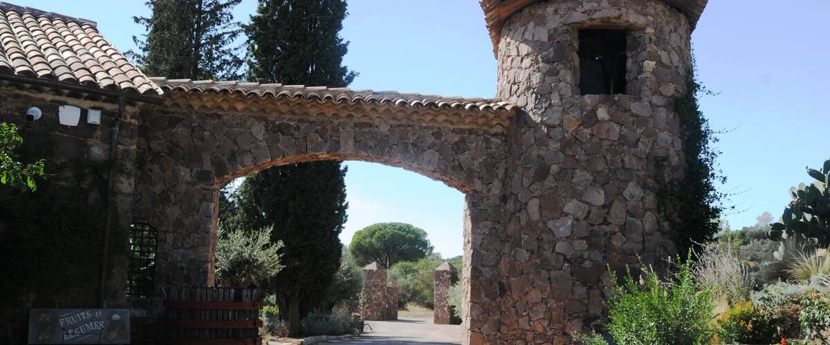 A stone archway leading to a tower with a tiled roof, surrounded by greenery and a sign indicating a fruit and legume stand.