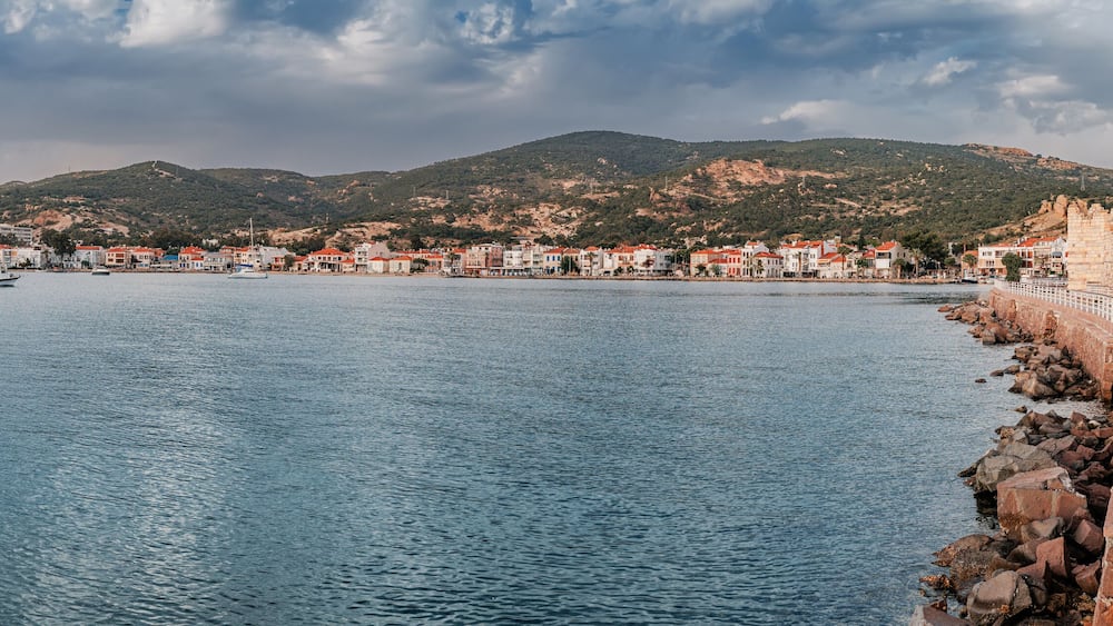 Coastal pathway beside calm waters, leading to a historic fortress in Foca with a Turkish flag, overlooking a charming town and hilly landscape