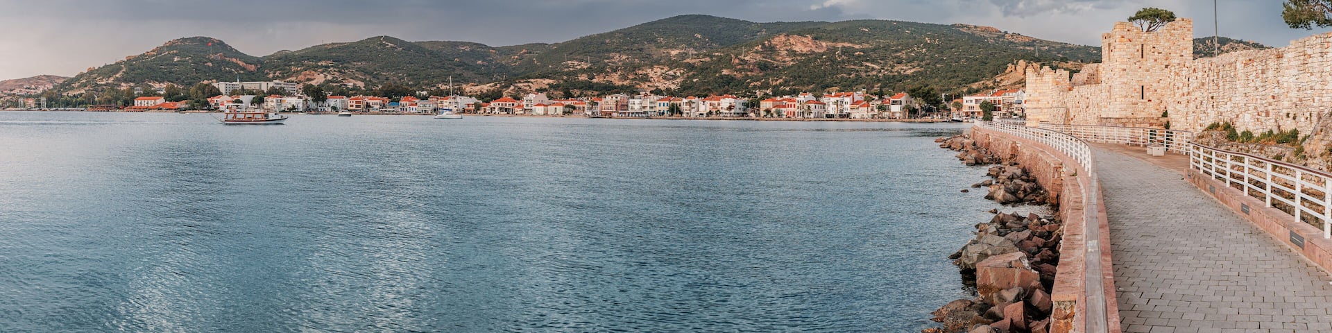 Coastal pathway beside calm waters, leading to a historic fortress in Foca with a Turkish flag, overlooking a charming town and hilly landscape