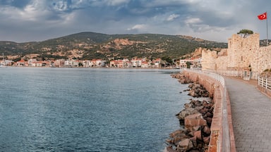 Coastal pathway beside calm waters, leading to a historic fortress in Foca with a Turkish flag, overlooking a charming town and hilly landscape