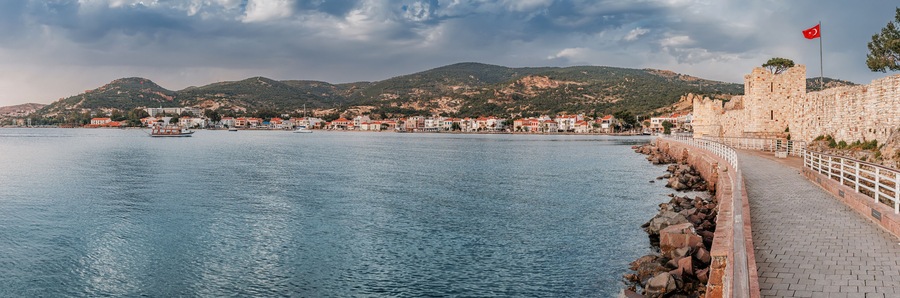 Coastal pathway beside calm waters, leading to a historic fortress in Foca with a Turkish flag, overlooking a charming town and hilly landscape