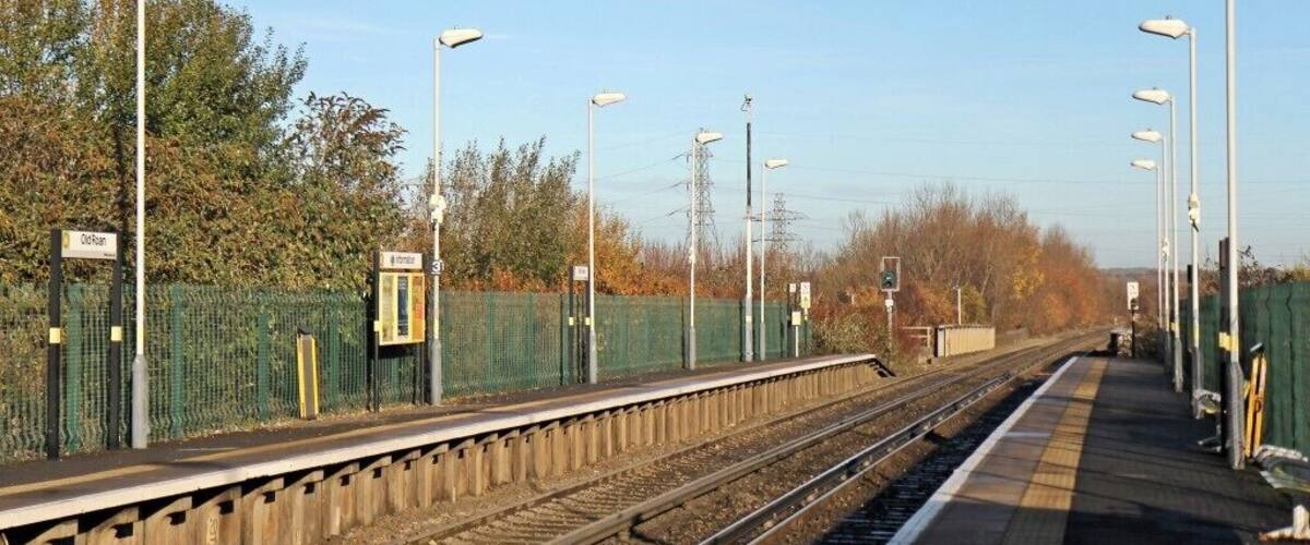 A railway platform with multiple tracks, overhead electric lines, street lamps, and a green fence.