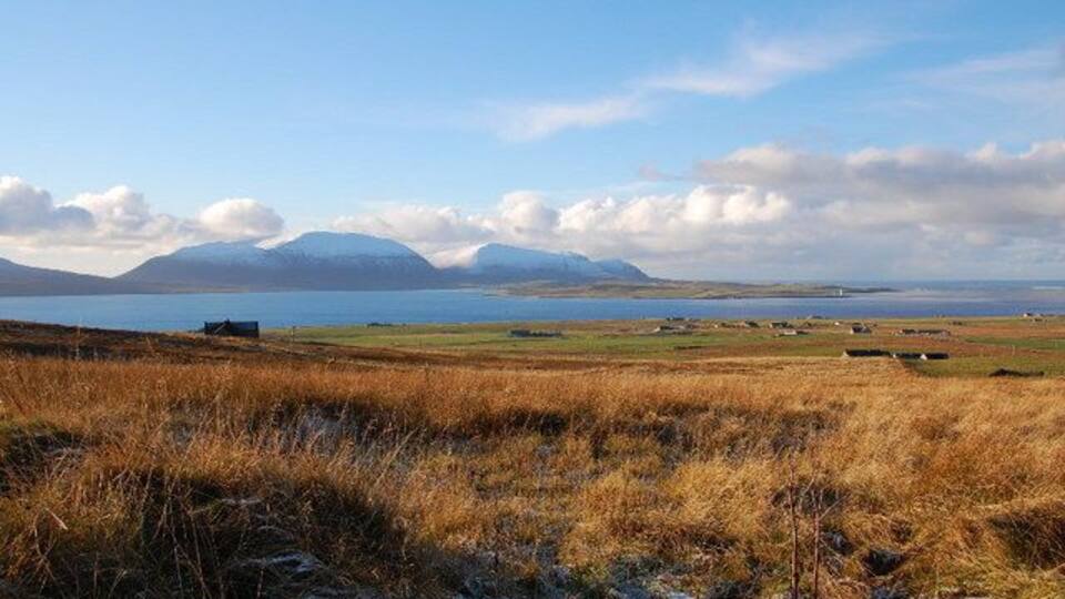 A coastal landscape with a grassy foreground, a body of water, and distant mountains under a clear sky.