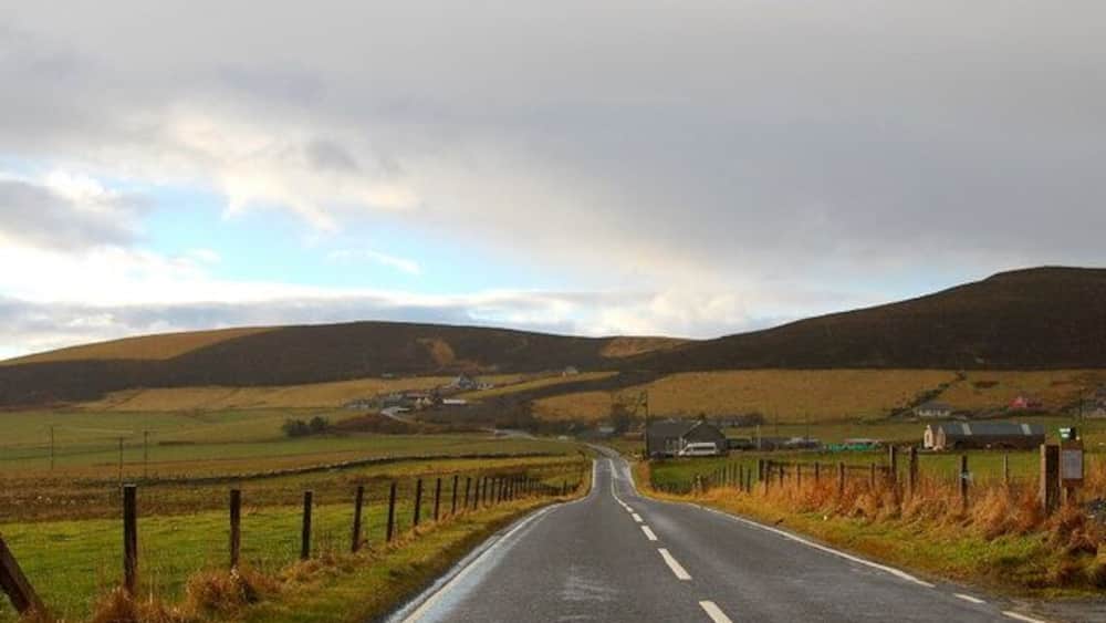 A rural road flanked by fields and hills under a cloudy sky.