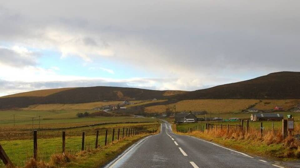 A rural road flanked by fields and hills under a cloudy sky.